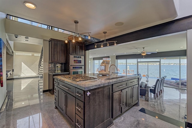 kitchen featuring a kitchen island with sink, granite finish floor, a water view, a sink, and dark brown cabinets