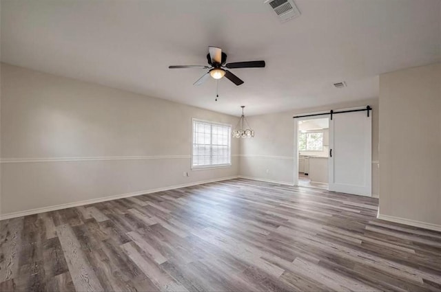 unfurnished room with a barn door, ceiling fan with notable chandelier, wood finished floors, visible vents, and baseboards