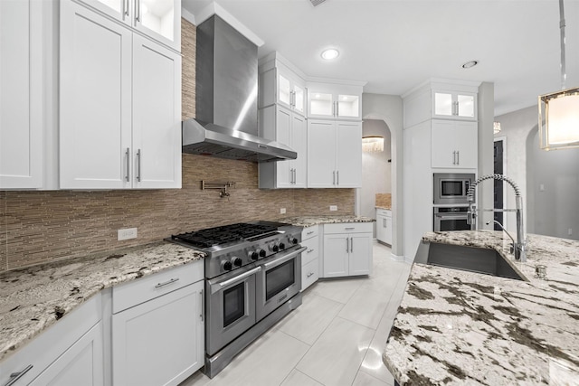 kitchen with stainless steel appliances, glass insert cabinets, white cabinetry, a sink, and wall chimney range hood