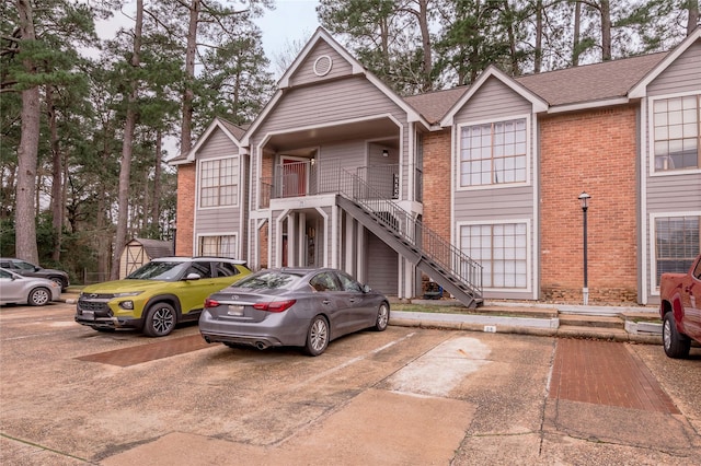 multi unit property with uncovered parking, brick siding, a shingled roof, and stairway