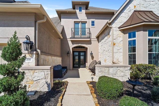 view of exterior entry with a balcony, a shingled roof, stone siding, french doors, and stucco siding