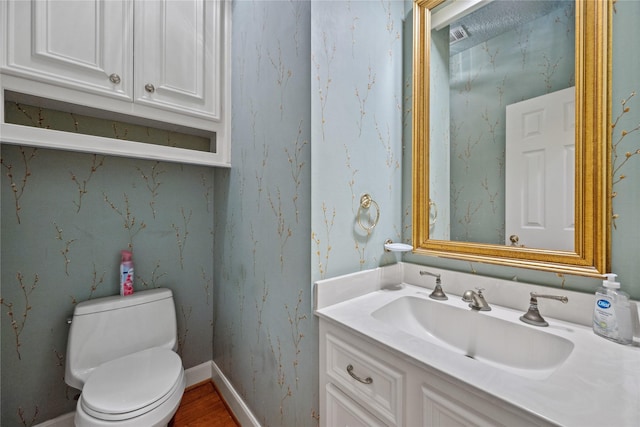 half bathroom featuring toilet, wallpapered walls, baseboards, and vanity