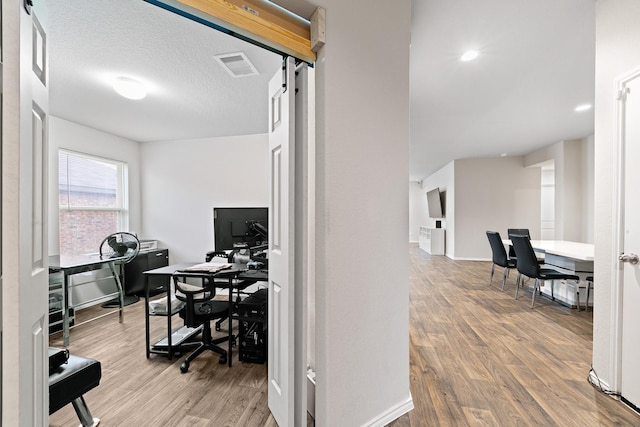 office space with a textured ceiling, a barn door, visible vents, baseboards, and light wood finished floors