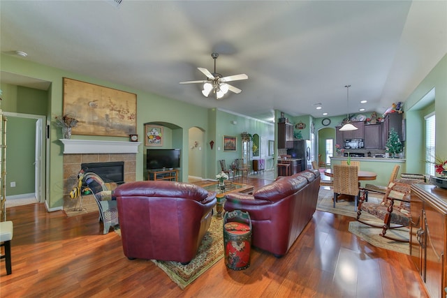 living room with baseboards, arched walkways, a ceiling fan, a tiled fireplace, and dark wood-style flooring