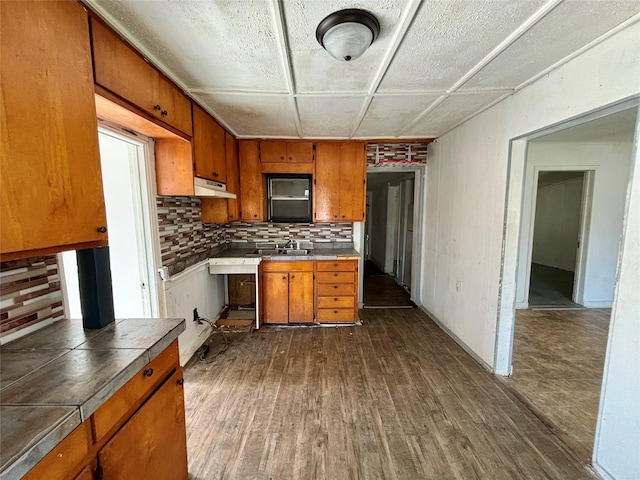 kitchen featuring under cabinet range hood, dark wood-type flooring, a sink, backsplash, and brown cabinets