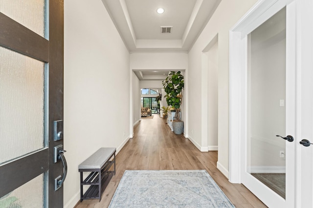 hall with a tray ceiling, light wood-style flooring, visible vents, and baseboards