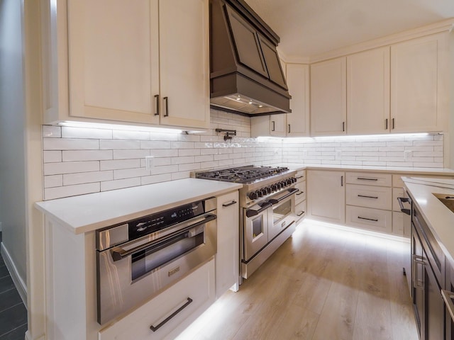 kitchen featuring light wood finished floors, custom exhaust hood, light countertops, decorative backsplash, and appliances with stainless steel finishes