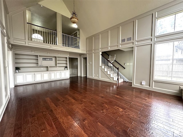 unfurnished living room featuring high vaulted ceiling, dark wood-style flooring, a decorative wall, and stairway