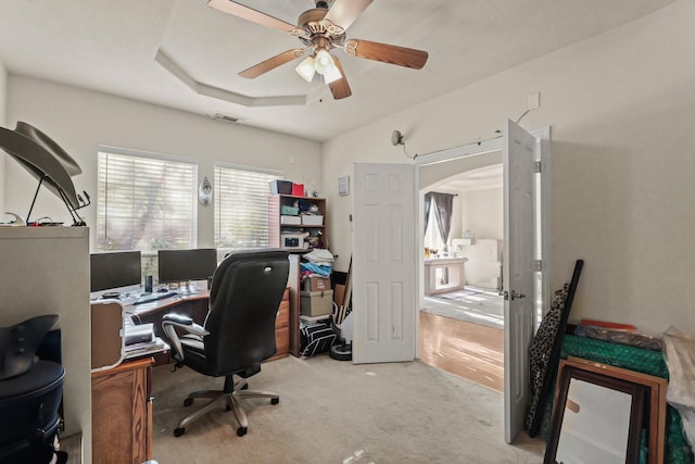 office space featuring arched walkways, light colored carpet, a ceiling fan, visible vents, and a raised ceiling