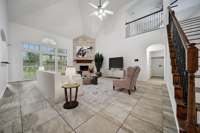 living area with arched walkways, high vaulted ceiling, a stone fireplace, a ceiling fan, and stairs