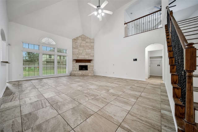 unfurnished living room with arched walkways, stairway, ceiling fan, a stone fireplace, and baseboards