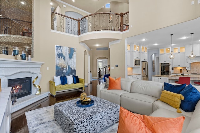 living area with arched walkways, dark wood-type flooring, a towering ceiling, stairs, and a glass covered fireplace