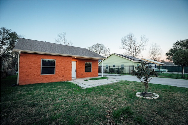 back of property at dusk with a patio, brick siding, fence, a lawn, and roof with shingles