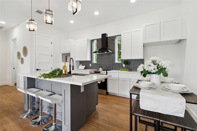 kitchen featuring stainless steel range with electric stovetop, a kitchen island with sink, light countertops, and wall chimney range hood