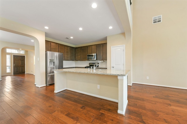 kitchen with arched walkways, light stone counters, appliances with stainless steel finishes, dark wood-style floors, and an island with sink