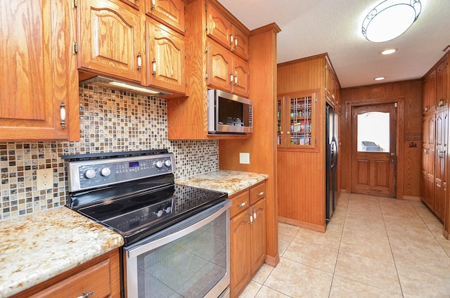 kitchen with light tile patterned floors, light stone countertops, stainless steel appliances, brown cabinets, and decorative backsplash