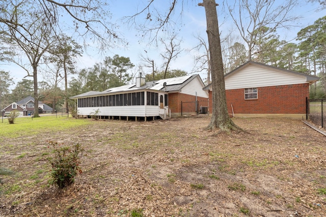 back of house featuring brick siding, fence, a sunroom, roof mounted solar panels, and a chimney