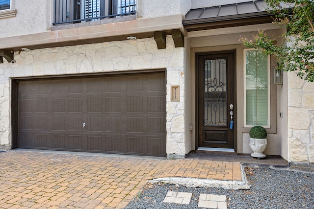 property entrance with metal roof, an attached garage, stone siding, stucco siding, and a standing seam roof