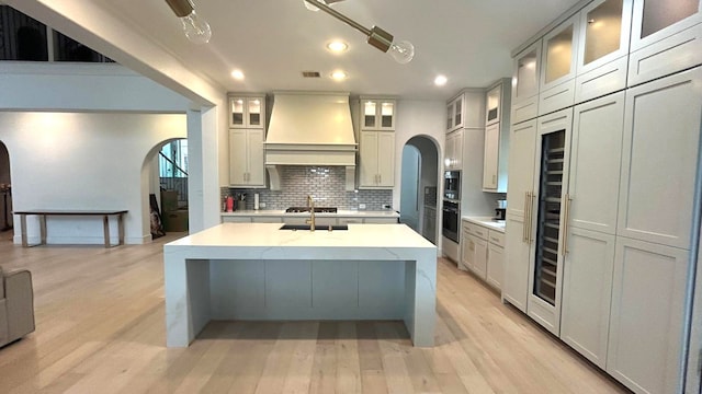 kitchen with arched walkways, stainless steel appliances, light countertops, custom exhaust hood, and glass insert cabinets