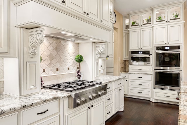 kitchen featuring light stone counters, premium range hood, appliances with stainless steel finishes, decorative backsplash, and glass insert cabinets