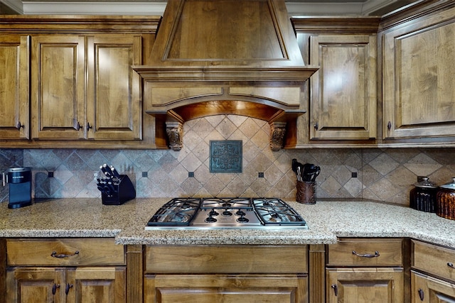 kitchen featuring brown cabinetry, light stone counters, premium range hood, stainless steel gas cooktop, and backsplash