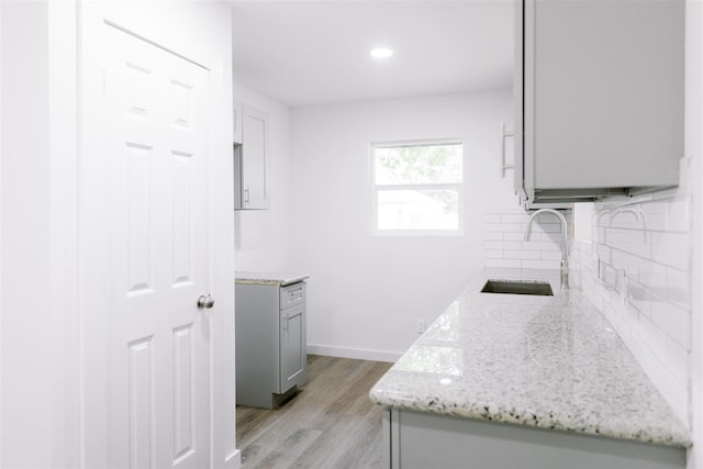 kitchen featuring light wood-style flooring, gray cabinetry, a sink, backsplash, and light stone countertops