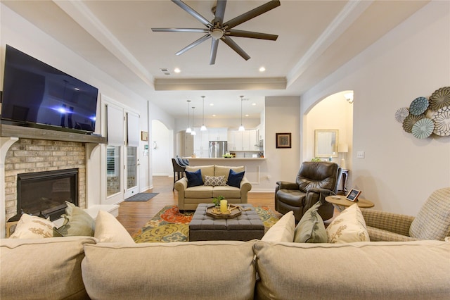 living area featuring arched walkways, a raised ceiling, a brick fireplace, and wood finished floors