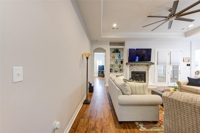 living area featuring arched walkways, dark wood-style flooring, baseboards, a brick fireplace, and a tray ceiling