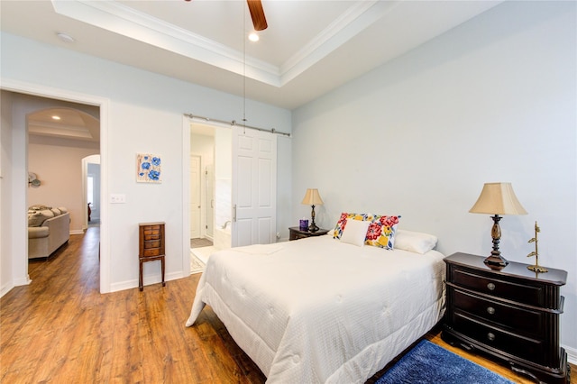 bedroom featuring a tray ceiling, arched walkways, a barn door, ornamental molding, and wood finished floors