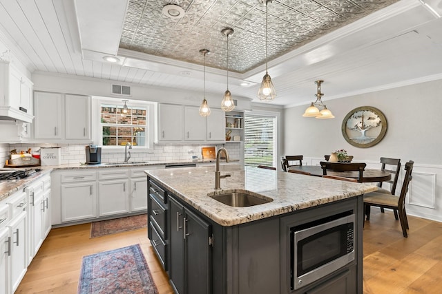 kitchen featuring decorative light fixtures, stainless steel microwave, white cabinets, a sink, and an island with sink