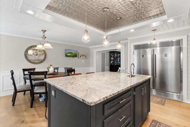 kitchen with built in refrigerator, a sink, light stone countertops, an island with sink, and an ornate ceiling