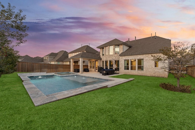 pool at dusk featuring a yard, an outdoor living space with a fireplace, a fenced backyard, and an in ground hot tub