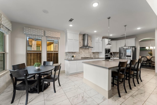 kitchen featuring an island with sink, wall chimney exhaust hood, decorative light fixtures, marble finish floor, and a sink