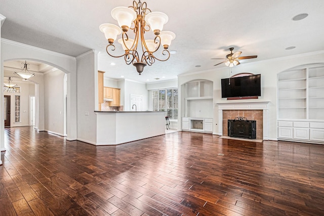 unfurnished living room featuring arched walkways, built in shelves, ceiling fan with notable chandelier, a fireplace, and wood finished floors