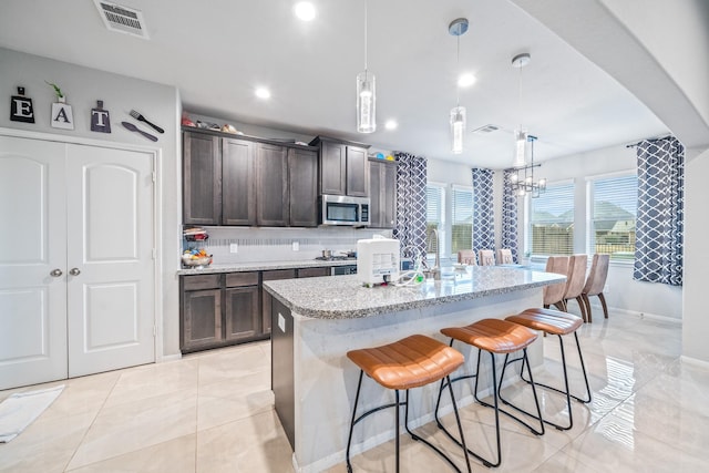 kitchen featuring light stone counters, a kitchen island with sink, visible vents, dark brown cabinets, and stainless steel microwave