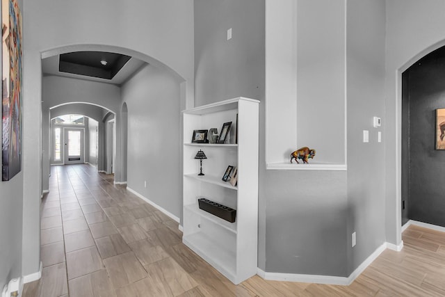 hallway with arched walkways, a tray ceiling, baseboards, and wood finished floors