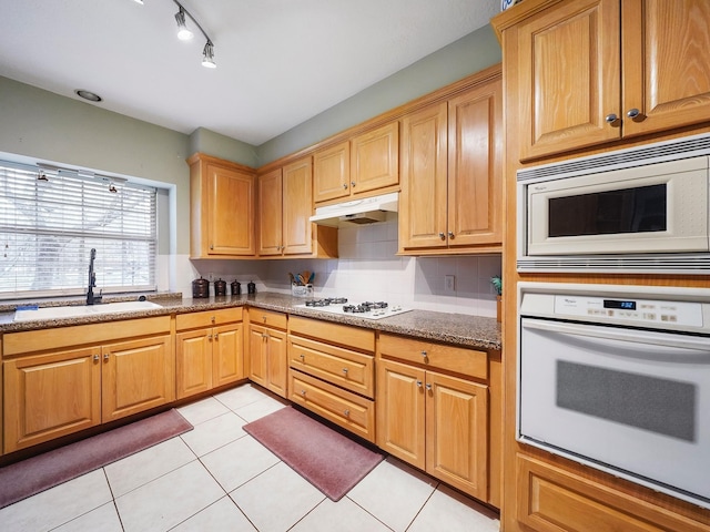 kitchen with light tile patterned floors, under cabinet range hood, white appliances, a sink, and dark stone countertops