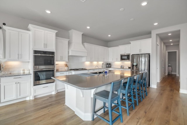 kitchen with light wood-style flooring, white cabinetry, appliances with stainless steel finishes, custom exhaust hood, and a center island with sink