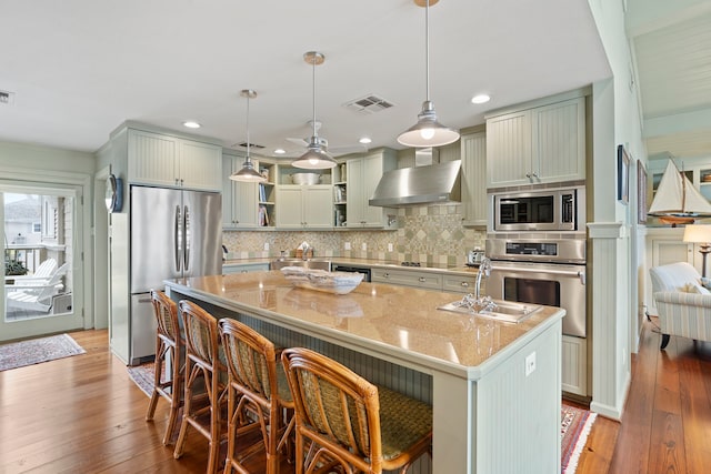 kitchen featuring light stone counters, stainless steel appliances, visible vents, wall chimney range hood, and an island with sink