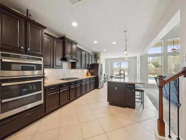 kitchen with light stone counters, pendant lighting, a center island with sink, a breakfast bar area, and stainless steel appliances