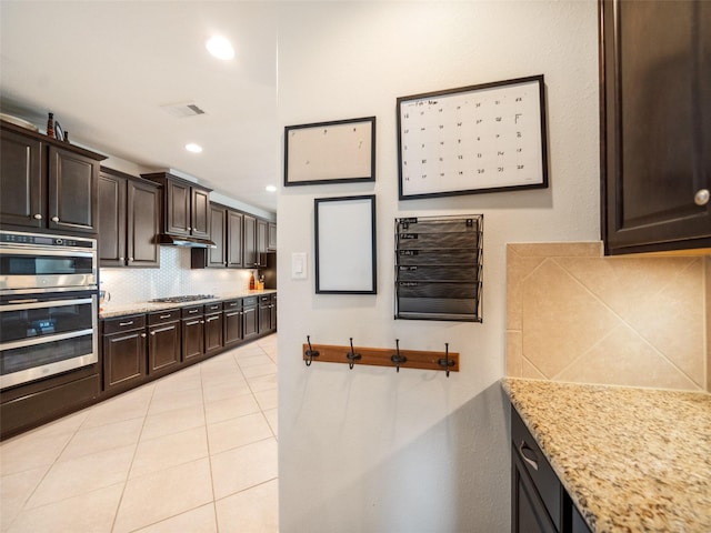 kitchen with light tile patterned floors, light stone counters, dark brown cabinetry, stainless steel appliances, and tasteful backsplash