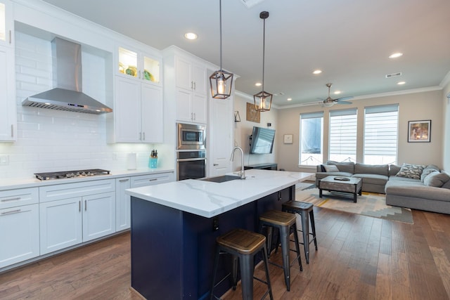 kitchen featuring stainless steel appliances, a sink, white cabinetry, open floor plan, and wall chimney exhaust hood