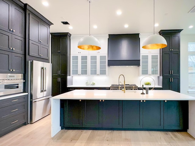 kitchen featuring stainless steel appliances, a kitchen island with sink, hanging light fixtures, and wall chimney exhaust hood