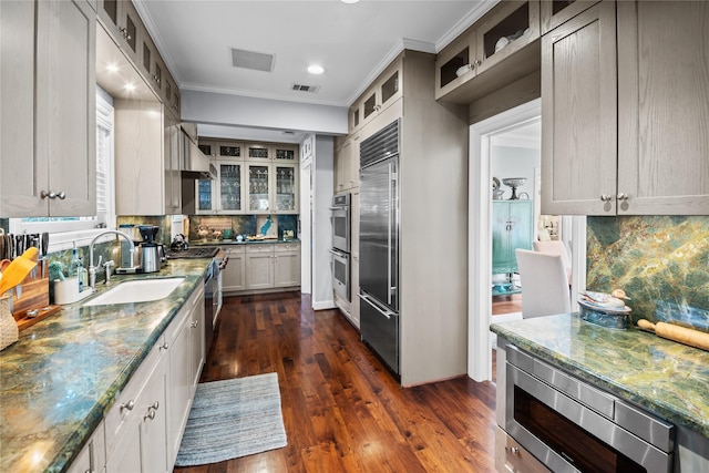 kitchen featuring tasteful backsplash, glass insert cabinets, a sink, light stone countertops, and built in appliances