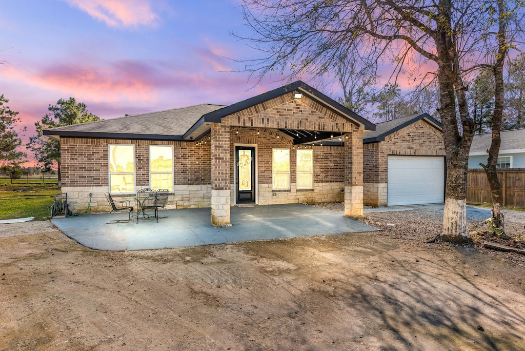 view of front of property featuring a garage, brick siding, a patio area, and a shingled roof