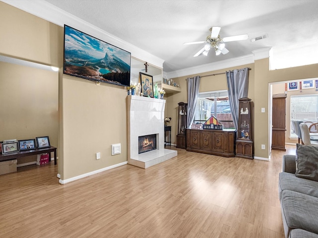 living room with baseboards, a ceiling fan, a tiled fireplace, ornamental molding, and light wood-style floors