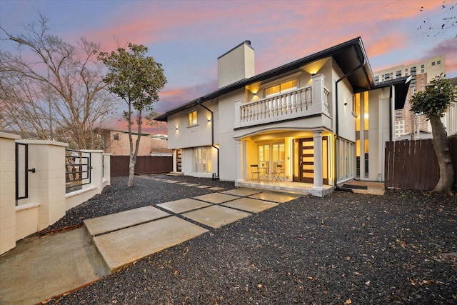 back of house at dusk featuring a balcony, fence, stucco siding, a chimney, and a patio area