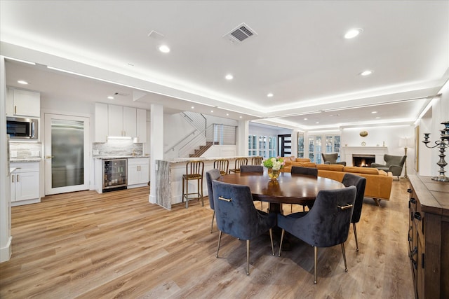 dining area with visible vents, light wood-style flooring, stairway, beverage cooler, and a warm lit fireplace