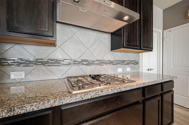 kitchen with stainless steel gas cooktop, exhaust hood, dark brown cabinets, backsplash, and light stone countertops