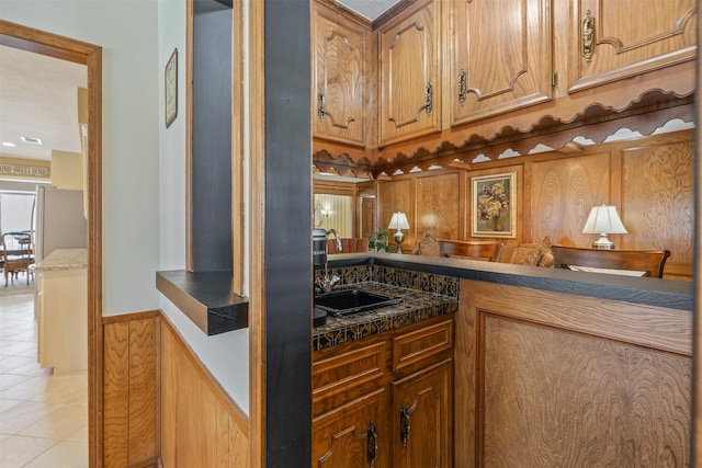 kitchen with light tile patterned floors, brown cabinetry, wainscoting, dark countertops, and a sink